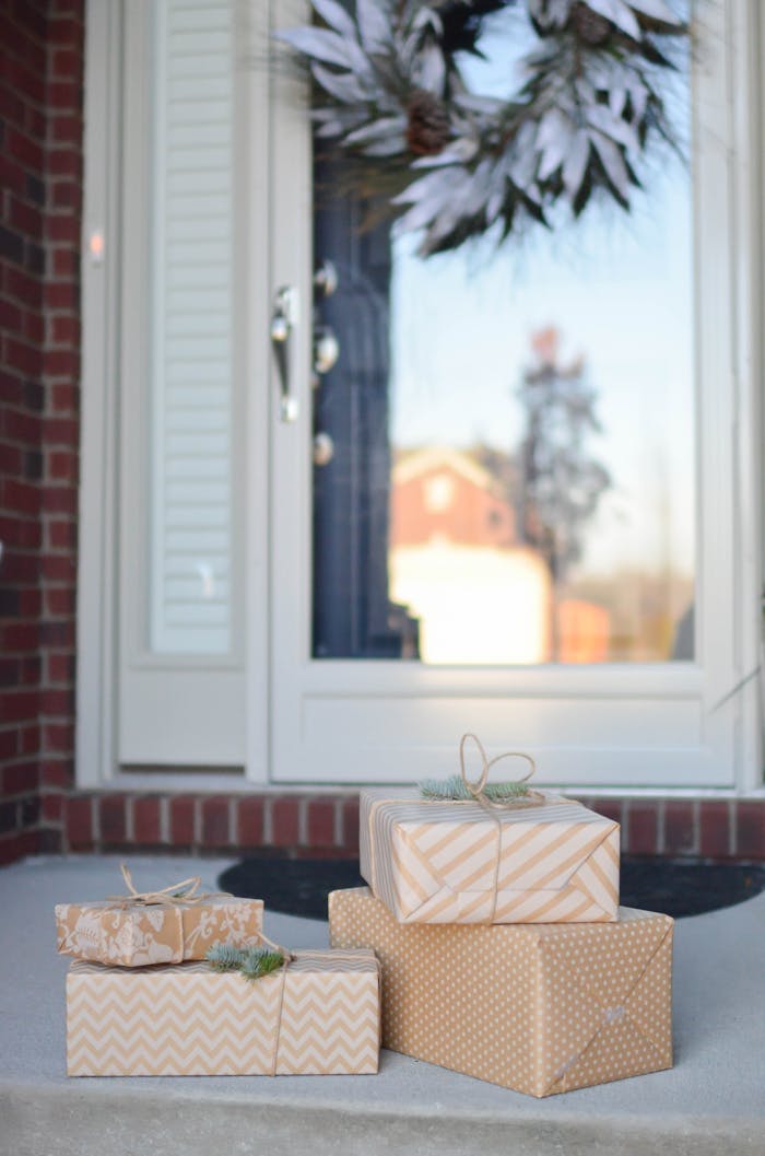 A stack of beautifully wrapped gifts sits on a doorstep with a reflective door and decorative wreath.